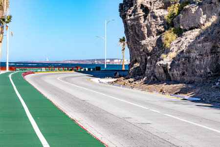 Bikeway along federal highway 11 towards del Ruso viewpoint, sea and industrial complex against blue sky background, rocky hillside at roadside, sunny day in La Paz, Baja Californiaの写真素材