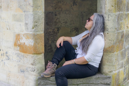 Pensive Latin American brunette woman sitting on edge of medieval waterhole taking a break, long gray hair wearing sunglasses, white casual clothing, enjoying a sunny day in South Limburg, Netherlandsの写真素材