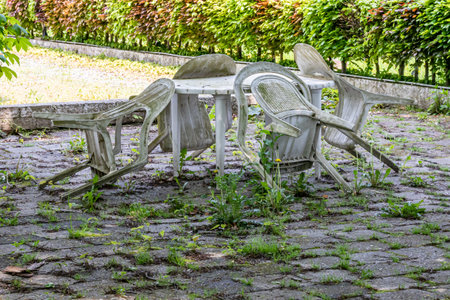 Old dirty old plastic chairs and table on a back garden terrace, dirt, weathered and covered with mold, unkempt floor with wild plants, sunny spring day, green bush in the backgroundの写真素材