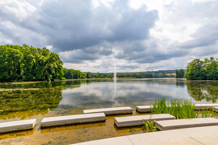 Concrete platforms on shore of Lake Echternach with waterjet gushing from a fountain, green leafy trees in background, reflection on water surface, panoramic landscape, summer cloudy day in Luxembourgの写真素材