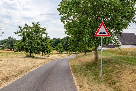 Country road with a traffic sign indicating speed bump, between dry grass, farmland and farms, green leafy trees in background, dry season due to climate change, summer day in Weidingen, Germanyの写真素材