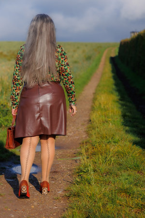 Rear view of elegant mature woman walking along a path in countryside, long straight gray hair, brown skirt, blouse with green touches, bag, stockings with back line and brown medium-heeled shoesの写真素材