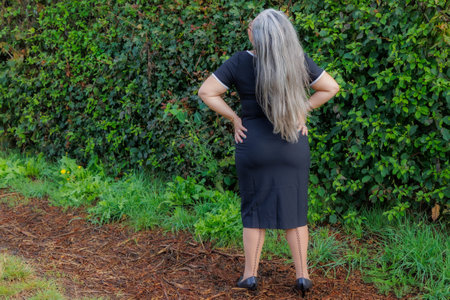 Rear view of full length mature woman in elegant black dress standing in garden, head tilted back,...