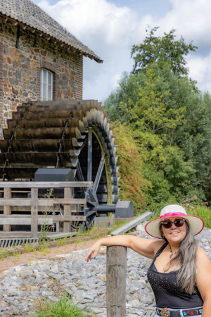 Happy smiling female tourist standing in front of old Eper or Wingbergermolen water mill, hat, sunglasses, tree on blurred background, sunny day in Terpoorten, Epen, South Limburg, Netherlandsの写真素材