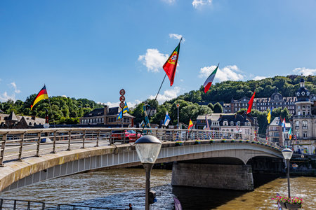 Vehicular bridge over Meuse river in Dinant city, waving flags, cars driving and cityscape with buildings and hill with leafy trees in background, sunny summer day in Namur province, Wallonia, Belgiumの写真素材