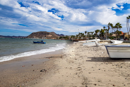 Coastal beach landscape, boats on sand and waters of Sea of ââCortez, palm trees on boardwalk and mountain against blue sky, day in La Paz, Baja California Sur Mexicoの写真素材