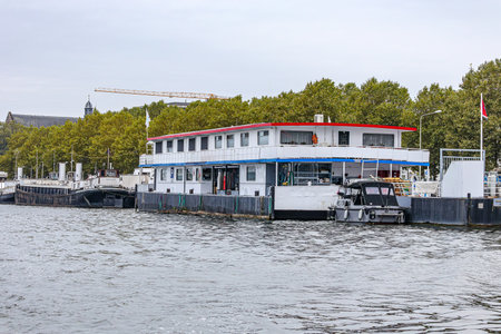 Group of boats moored at embankment quay along river Meuse, green foliage of trees in background against gray sky, calm water, Maastricht city in South Limburg, Netherlandsの写真素材