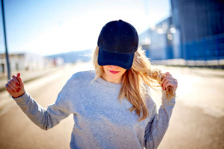 Girl with a cap and long blonde hair, wearing a grey sweatshirt, walking down the street in an empty urban area on a very sunny day and playing with her hairの写真素材