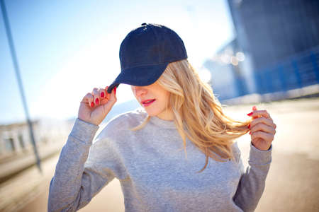 Girl with a cap and long blonde hair, wearing a grey sweatshirt, walking down the street in an empty...