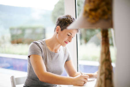 Woman sitting at her desk by a large window inside her house reading with concentration and writing in her diaryの写真素材
