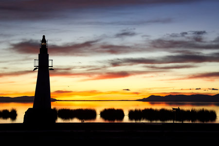 Spectacular sunset on a calm water lake with an incredibly colorful sky and a small lighthouse in the foreground and a boat with a fisherman among some reedsの写真素材