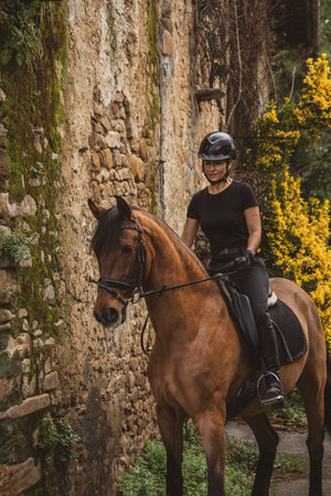 Female rider dressed in black with her brown horse riding through a village with stone wallsの写真素材