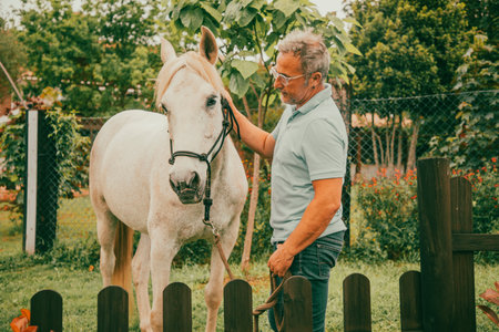 Man with white hair and glasses in blue jeans and blue polo shirt walking with a white horse through the fieldsの写真素材