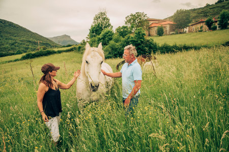 Man with white hair and glasses in blue jeans and blue polo shirt walking with a white horse through the fieldsの写真素材