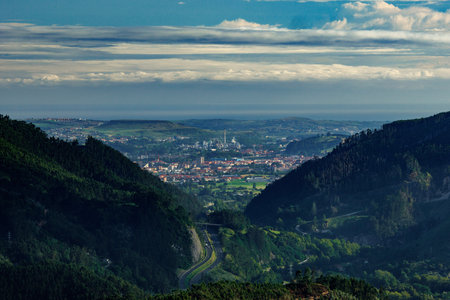 The image offers a breathtaking view of a city nestled between lush, green hills with a distant horizon blending into the sky. The foreground is dominated by densely forested hills, which frame the valley where the city lies, creating a natural corridor leading the viewer's eyes towards the urban landscape. The cityscape itself is composed of various buildings, roads, and structures, with a mix of industrial and residential areas, showing a vibrant mix of life and activity. Beyond the city, rolling hills extend into the distance, eventually meeting the calm and expansive sea under a hazy horizon. The sky above is partly cloudy, with soft layers of clouds that add depth and drama to the scene. This composition beautifully captures the contrast between the untouched natural landscape and human-made developments, evoking a sense of harmony between nature and civilization.の写真素材