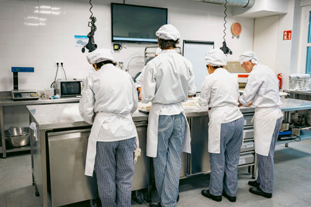 Group of pastry students in a professional bakery kitchen, dressed in white uniforms, working on dough preparation. Culinary education and teamwork in a modern food lab.の写真素材