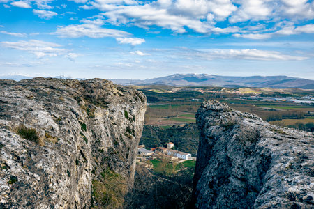 View from high rocky cliffs looking down at a small rural village, surrounded by farmlands and distant mountains under a blue sky with scattered clouds.の写真素材