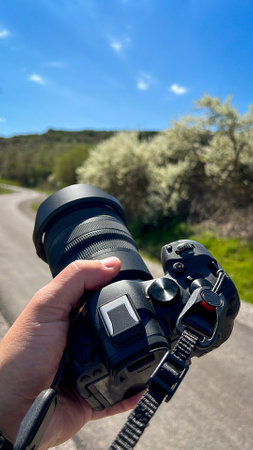 Close-up of a person holding a professional camera outdoors, ready to shoot on a sunny day, with a scenic rural road and blooming nature in the background.の写真素材