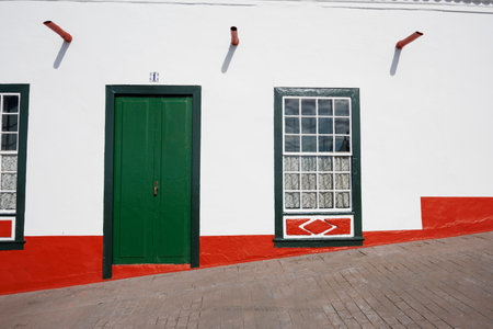 Geometric architectural detail of a bright white wall with bold colors, traditional door and window, captured on an inclined paved street.の写真素材