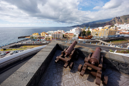 Old artillery pieces aligned on a fortress terrace overlooking the sea and urban skyline, with dynamic clouds in the background.の写真素材