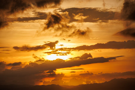 Dramatic sky with layered clouds illuminated by warm orange and yellow hues as the sun sets behind the distant hills and oceanの写真素材