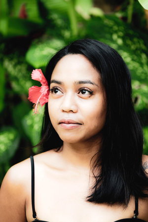 Natural beauty portrait of a woman posing among vibrant green leaves with a red flower tucked behind...