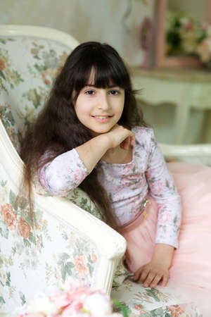 A girl in a pink dress sits on a vintage sofa surrounded by flowers.の写真素材