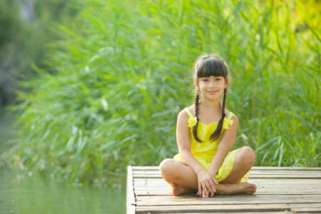 Child girl in a bright yellow dress and barefoot sitting on the pier on the shore of the pond.の写真素材
