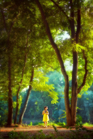 The baby girl in a bright yellow dress stands under the big tall trees by the river.の写真素材