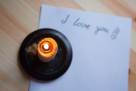 The inscription I love you in the form of a letter by hand on white paper and a candle burning on...