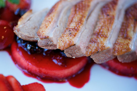 Pieces of roast duck meat lie on a white plate. Under the meat is a red sweet sauce. Decorated with vegetables and fruits. The concept of recipes from the restaurant and dishes from the chef.の写真素材