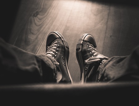 First-person perspective on two legs wearing retro sneakers and jeans while sitting, wooden floor, moody light and shadows, vintage look - landscape formatの写真素材