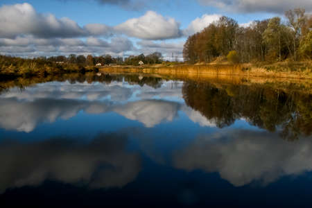 View on autumn landscape of river and trees in sunny day. Forest on river coast in autumn day. Reflection of autumn trees in water. Autumn in Latvia. Autumn landscape with colorful trees, yellow grass and river.の写真素材