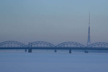 Railway bridge and television tower in winter. Railway bridge over the frozen river Daugava in Riga.TV tower and railway bridge on blue sky background in winter. Riga view in winter.の写真素材