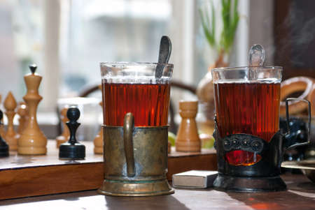 Two glasses of tea in old cupholders on a wooden table next to the chessboard near the window