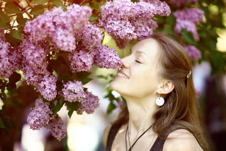 Beautiful young woman enjoying the smell of lilac in blossom garden in sunny spring day. Pretty female and flowers. Aromatherapy and springtime conceptの写真素材