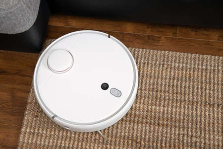 Robotic vacuum cleaner runs under sofa in room on laminate floor modern smart cleaning technology housekeeping.の写真素材