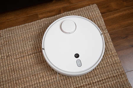 White robotic vacuum cleaner on laminate floor cleaning dust in living room interior. Smart electronic housekeeping technology.の写真素材