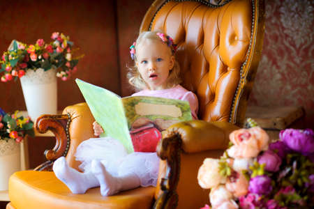 beautiful little girl, sits in a chair with the book in hands, with an amazed lookの写真素材