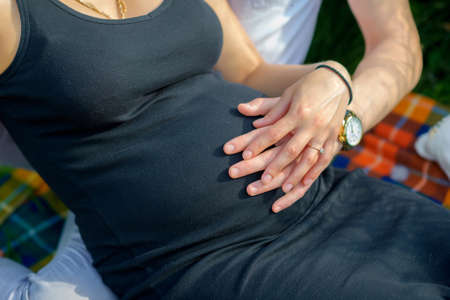 pregnant woman outdoors, sits on a grass, the husband gently embraces a tummy a hand, happy...