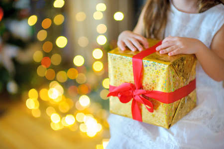girl sits near a fir-tree and holds in hand a Christmas gift, bright multi-colored sparks on a background, expectation of a holidayの写真素材