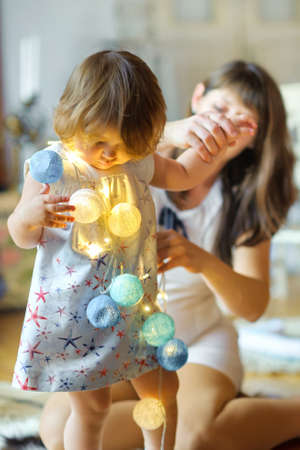 Mother together in the little daughter, sit on a floor near a festive fir-tree, play a garland,...