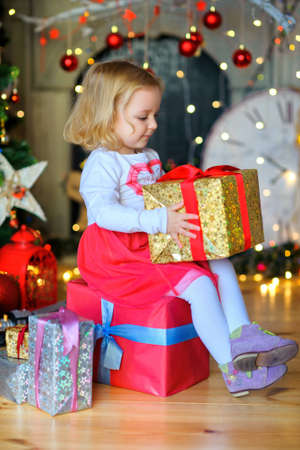 happy little girl, sits on a gift box near a festive fir-tree, holds gifts in hand. Waiting for a...