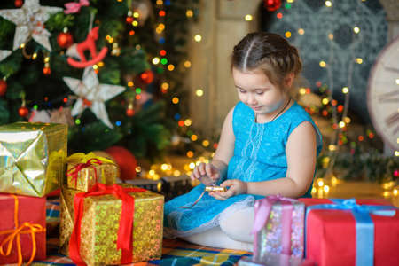 happy little girl sits on a floor near a festive fir-tree and opens gifts. On a background the Christmas tree. Waiting for a holiday.の写真素材