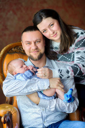 Happy family with the newborn son, sit on a chair the father carefully holds the baby on hands....