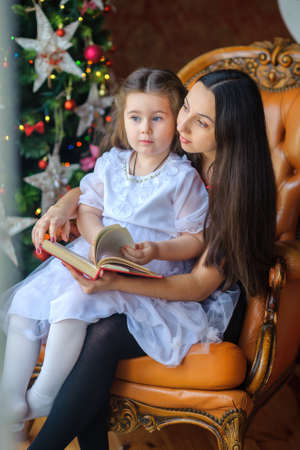 Mother with the beloved little daughter sits on a chair and reads fairy tales. Gently embraces and smiles. Nazadny plan festive fir-trees.の写真素材