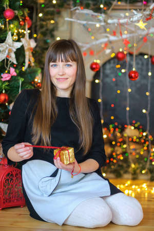 beautiful girl sidiit on a floor near a festive fir-tree, holds a gift in hand and gently smiles.のeditorial素材