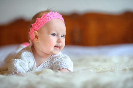 the newborn baby with big beautiful eyes and a knitted pink crown on the head, lies on a sofa with curiosity looks aroundの写真素材