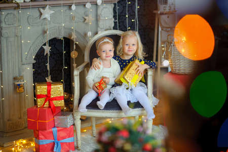 two beautiful sisters sit on a chair in an interior of studio, with gift boxes in hands, embrace and...