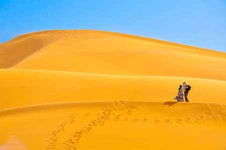 couple walks on high sandy dunes in bright sunny day, leave behind marks on sand, wind develops the girl's dressの写真素材